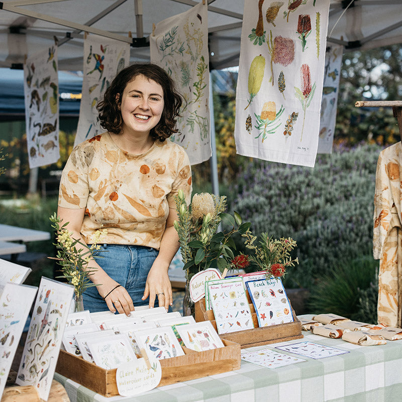 Artist Claire Mosley standing at her market stall in Melbourne, Victoria. She is surrounded by her illustrated products.