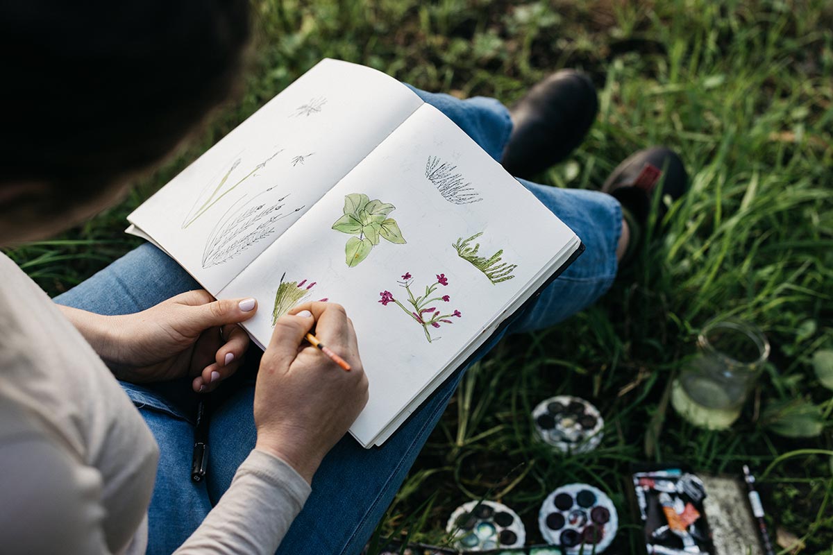 A photo of a person sitting outdoors, painting flowers and leaves with watercolour paints in an open notebook