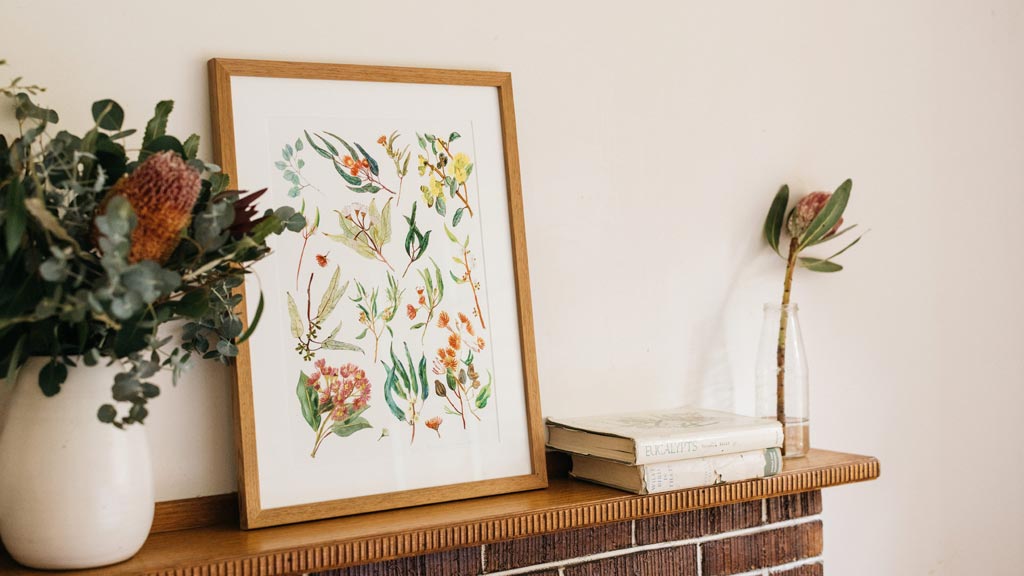 A mantlepiece featuring a vase of Australian native flowers, a framed illustration of native australian flowers, and some books
